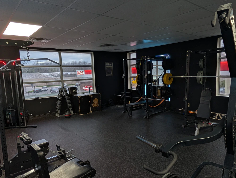 Front training floor at Retraine with squat racks, dumbbell tower, and Bells of Steel bench under window light.