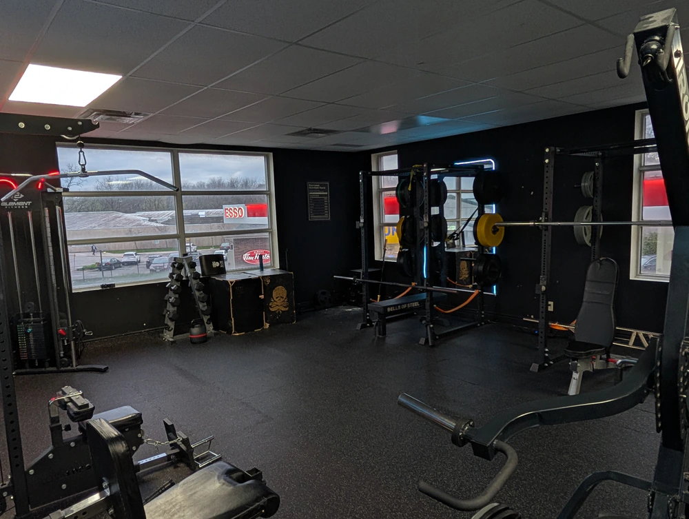 Front training floor at Retraine in Pickering with cable station, dumbbell tower, bench and squat rack under window light.